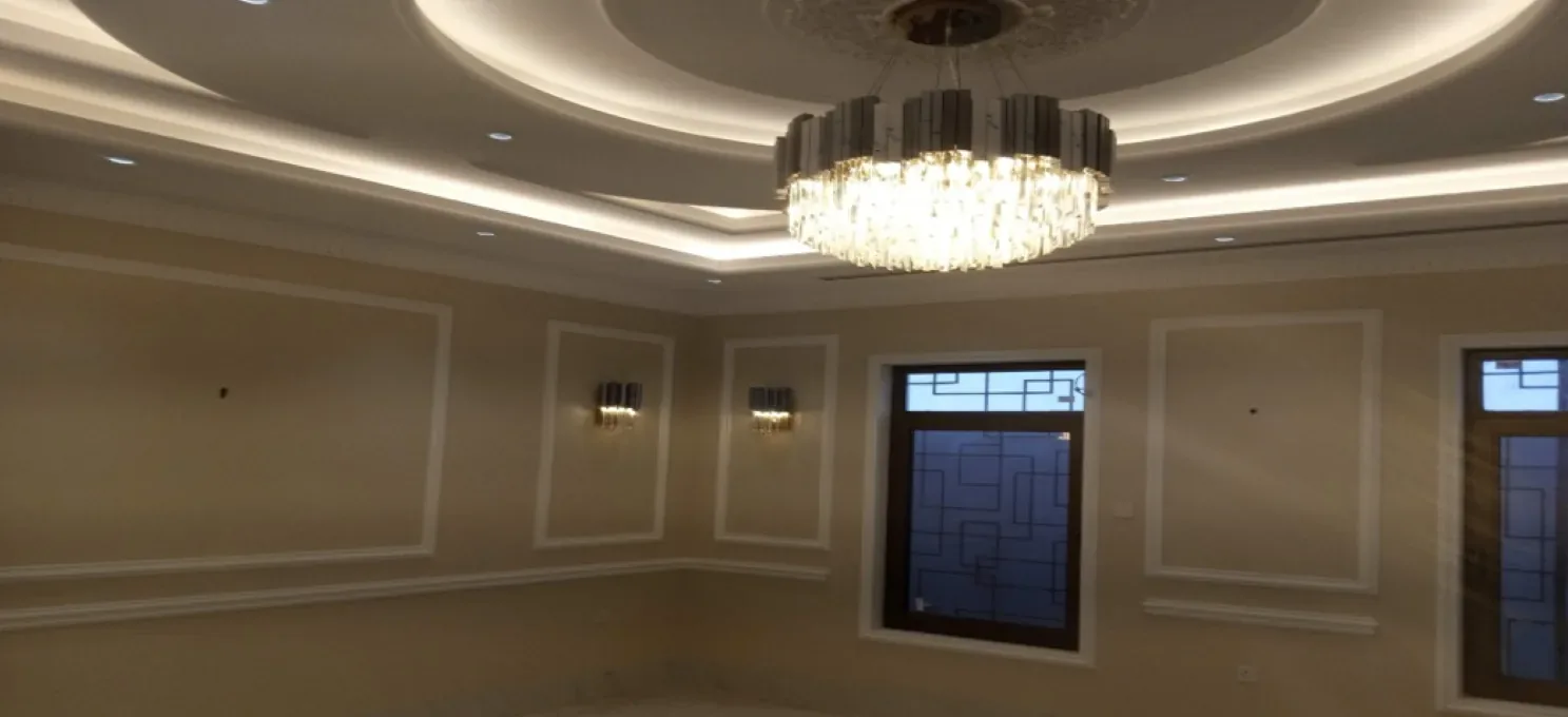 Cream room with a cove-lit tiered ceiling, crystal chandelier, wall lights and dark-framed windows with geometric grille patterns.