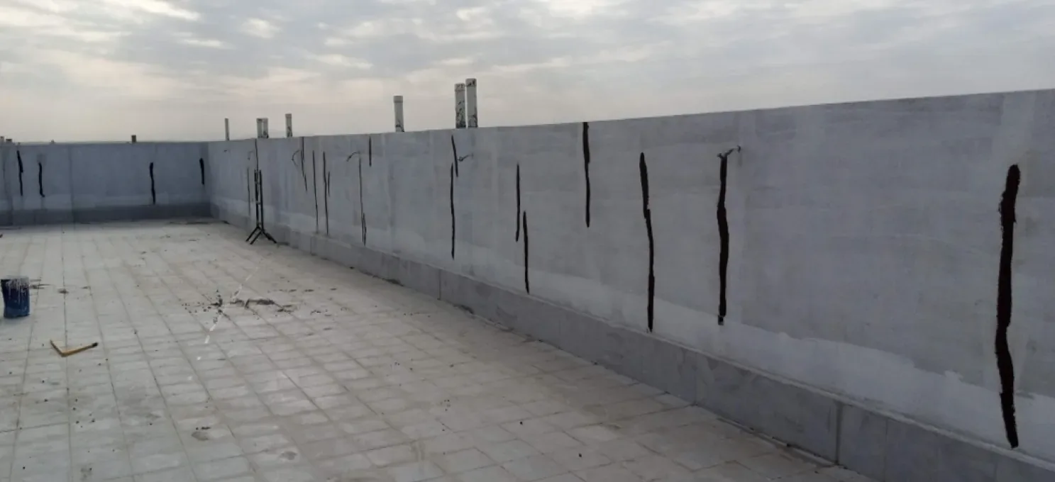 Tiled roof floor beside a grey parapet wall with vertical black bituminous strips; a blue bucket at left, cloudy sky.
