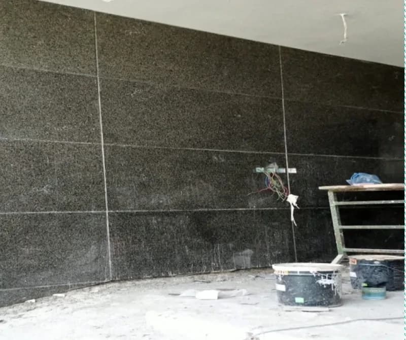 Dark granite-clad wall with wire ends near a slab joint, a scaffold trestle and paint tubs standing on dusty concrete.