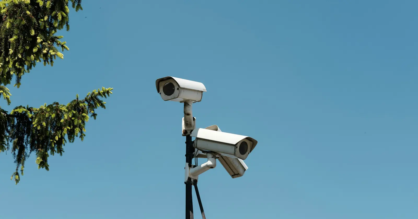Two white bullet-style CCTV cameras mounted on a steel post against a clear blue sky