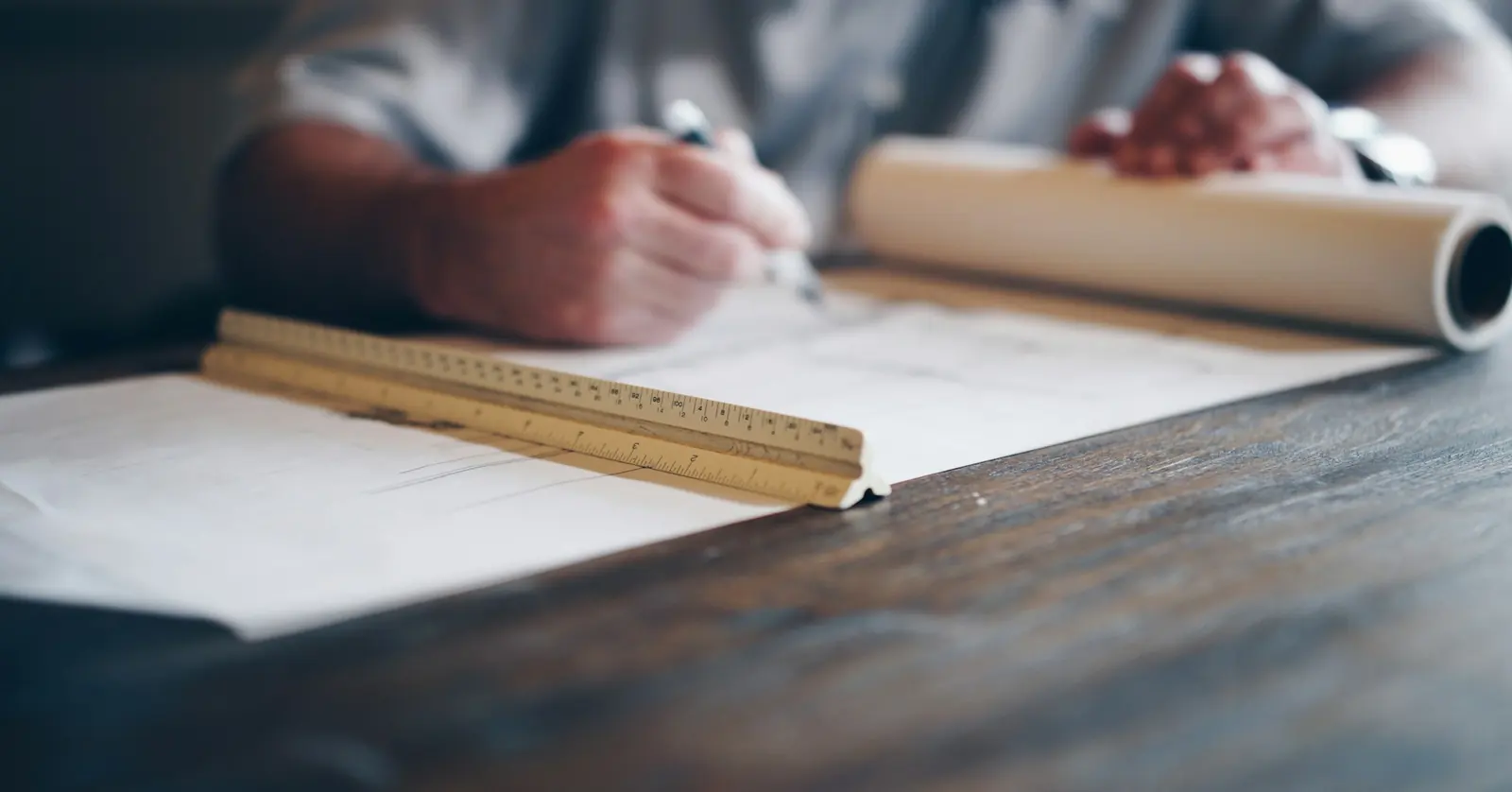 A hand marking up drawings with a scale ruler beside a roll of plans on a wooden desk