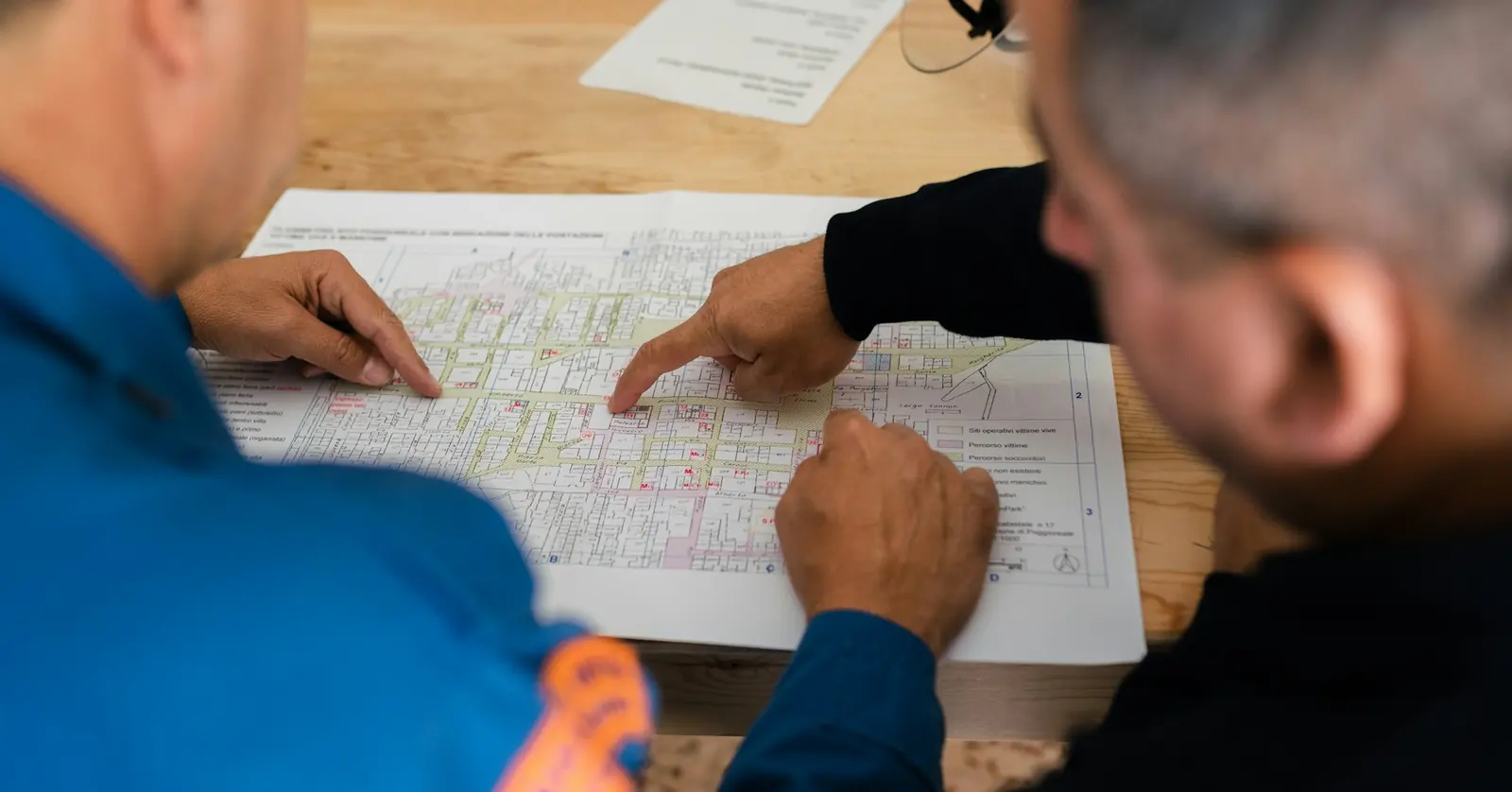Two men pointing at details on a printed plan laid on a wooden table