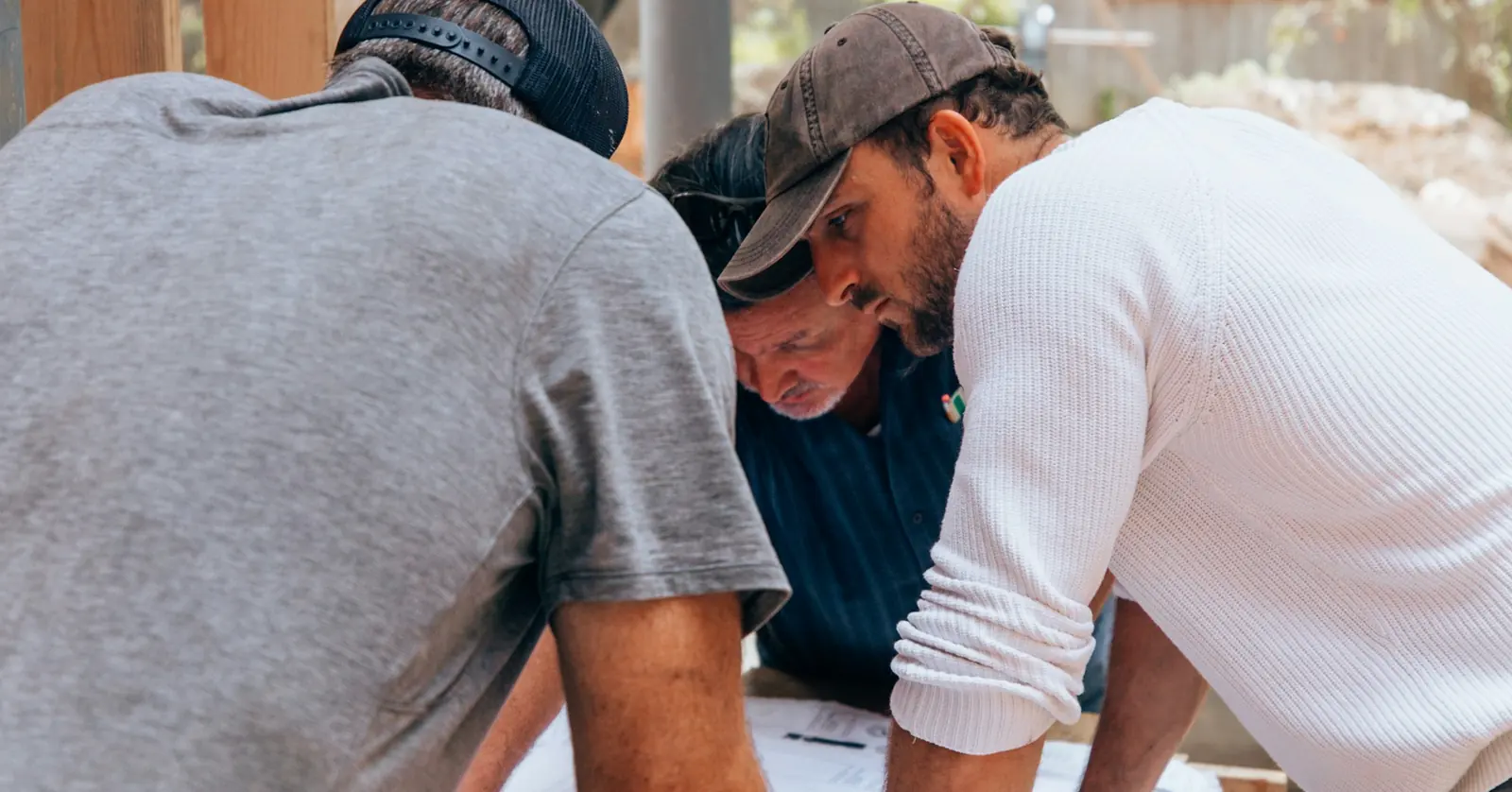 Three men bending over drawings on a makeshift table at a building site