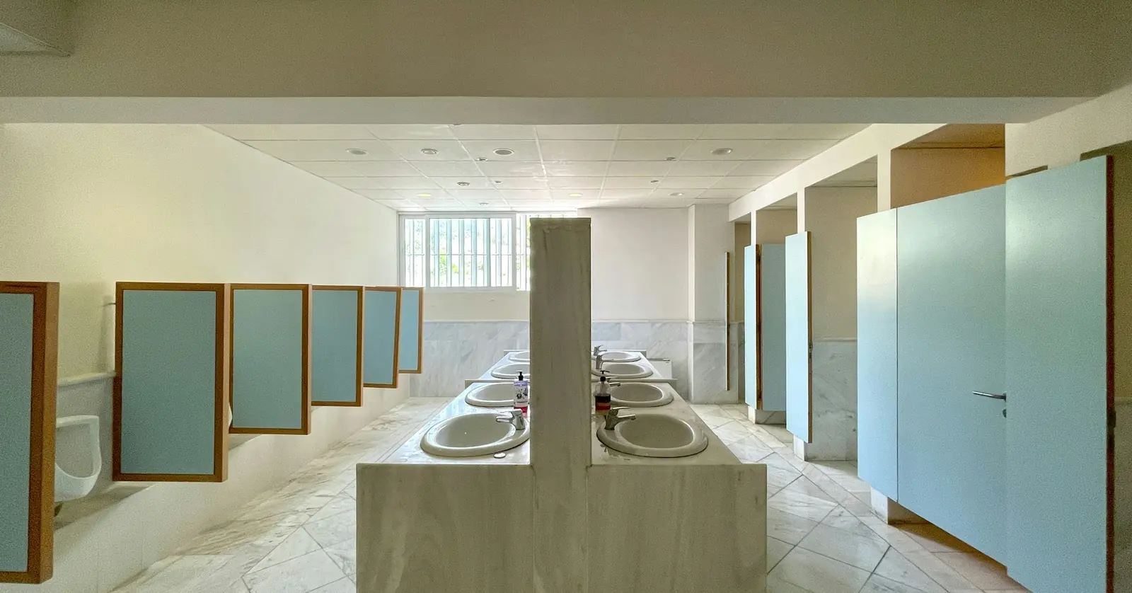 Public restroom with a row of pale-green cubicle partitions and doors beside a long stone-topped washbasin counter