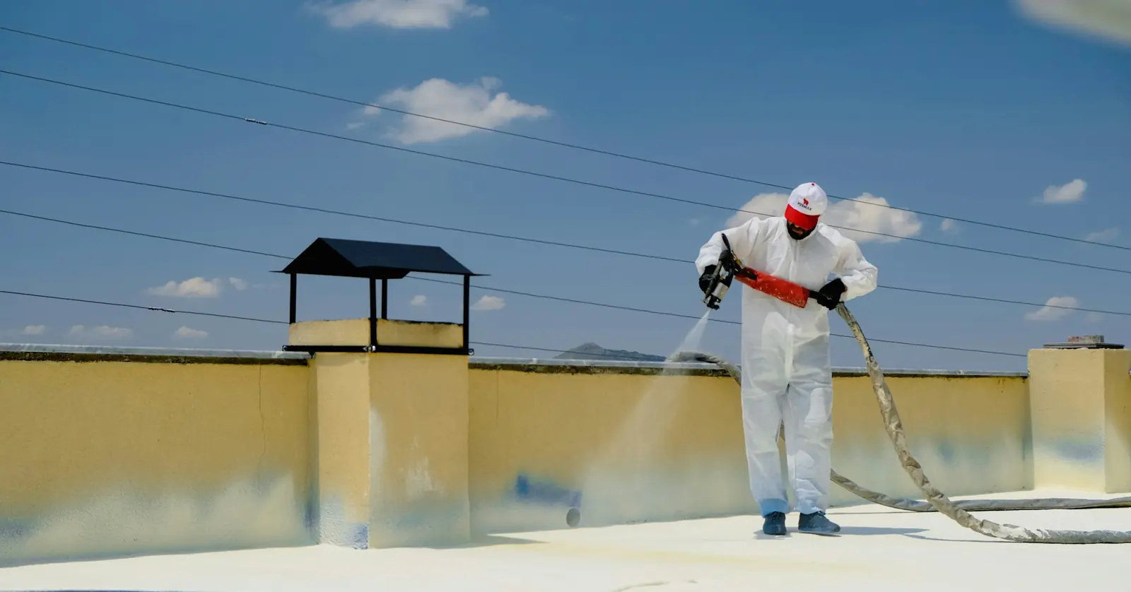 Worker in a white protective suit spray-applying a light coating over a flat roof beside a parapet wall