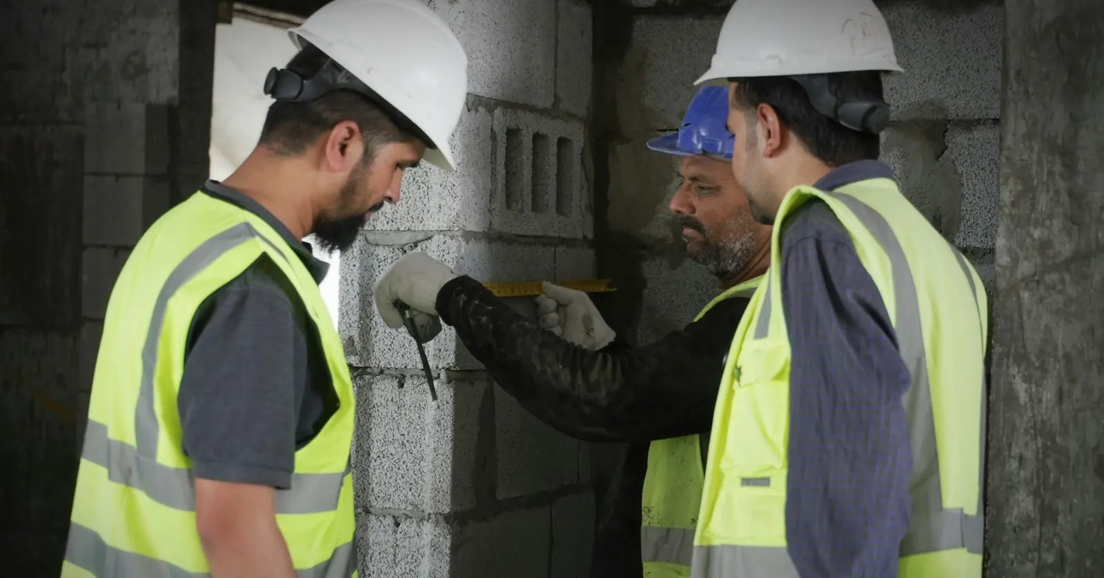 Three site staff in hard hats and high-visibility vests checking a concrete block wall with a measuring tool