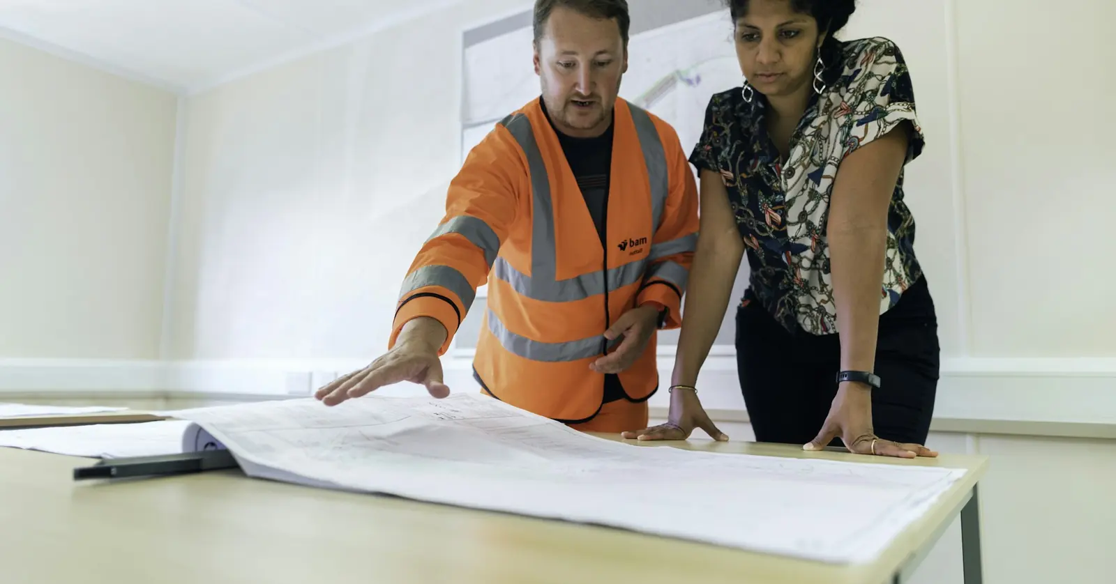 Two colleagues in a site office leaning over large drawings on a table